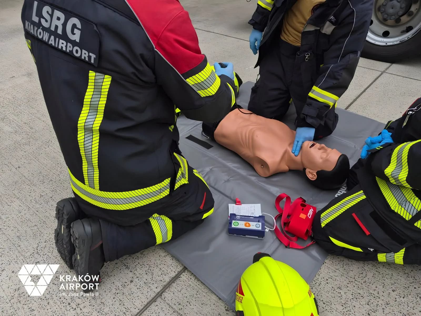 Dwaj strażacy Lotniskowej Służby Ratowniczo-Gaśniczej Kraków Airport klęczą przy fantomie na płycie lotniska, obok defibrylator ViVest Pocket AED P3