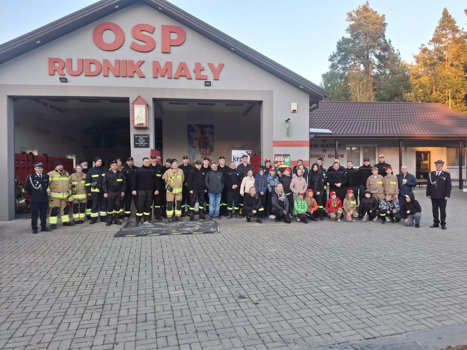 Druhowie OSP, członkowie młodzieżowej drużyny pożarniczej i mieszkańcy przed remizą w Rudniku Małym, z szafką AED na ścianie
